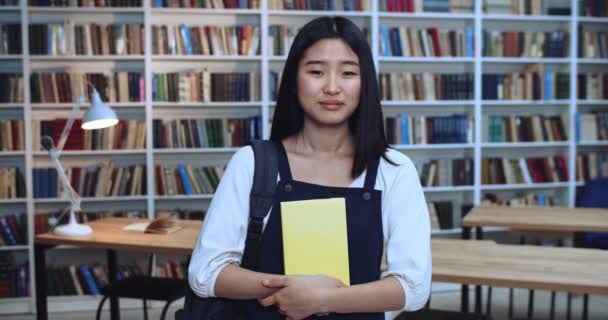 Porträt eines süßen asiatischen Mädchens mit schönen natürlichen Haaren, das ein gelbes Buch hält und in der Bibliothek studiert. Bücherregal im Hintergrund. — Stockvideo