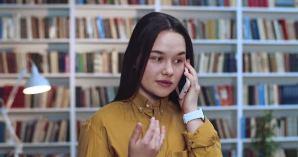 Retrato de una bonita estudiante geek bellísima en voz alta y emocionalmente hablando por teléfono y haciendo un montón de gestos por sus manos en la biblioteca . — Vídeos de Stock