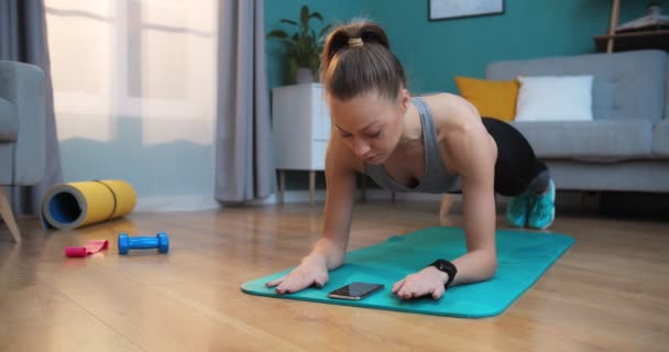 Starkes schönes Fitness-Girl in grauer Sportbekleidung, das Brettübungen macht, während es eine Stoppuhr am Telefon bedient. Frauentraining zu Hause im Wohnzimmer mit modernem Interieur. Aktive Frauen treiben Sport — Stockvideo