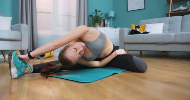 Atleta femenina en posición lateral estirándose sobre una alfombra azul en el suelo de la sala de estar mientras el gato duerme en el fondo. Deporte y fitness. Entrenamiento, concepto de entrenamiento. Chica estiramiento activo haciendo deportes . — Vídeos de Stock