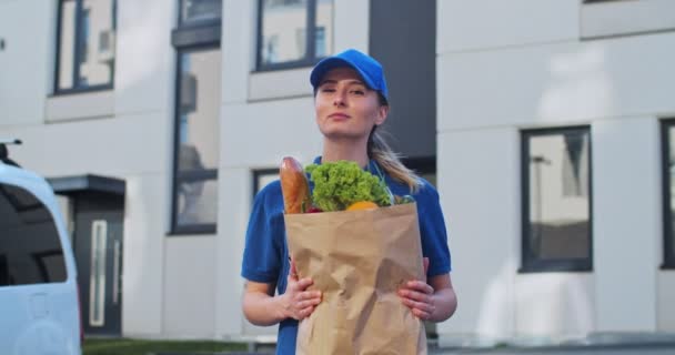 Portret van vrolijke jonge blanke aantrekkelijke bevallingswerkster met pet in pakje met vers eten in de hand en glimlachend naar de camera buiten. Vrouwelijke koerier met tas van goederen in tijden van pandemie. — Stockvideo