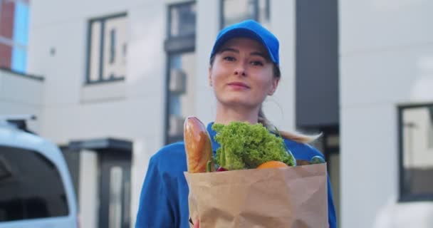 Ritratto di bella donna felice caucasica in uniforme blu e cappuccio dalla consegna del servizio girando e sorridendo alla macchina fotografica con pacchetto di cibo biologico. Carino corriere femminile all'aperto con verdure . — Video Stock