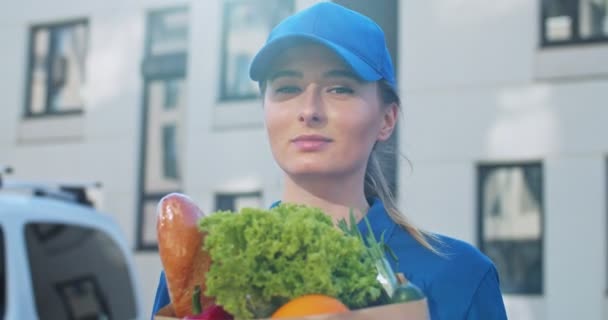 Gros plan de belle jeune femme caucasienne en bonnet bleu souriant joyeusement à la caméra avec sac de nourriture en plein air. Courier de livraison femelle tenant paquet avec des légumes regardant la caméra avec le sourire . — Video