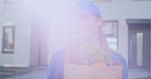 Portrait d'une jolie jeune femme caucasienne heureuse au chapeau bleu et uniforme souriant à la caméra avec sac de nourriture fraîche au soleil. Gros plan du paquet de courrier féminin avec épicerie en plein air . — Video