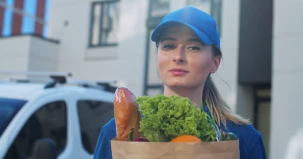 Portrét krásně usměvavé bělošské dívky v modré čepici, radostně hledící na kameru s pytlíkem čerstvého jídla. Detailní záběr kurýra držícího balíček s potravinami venku. — Stock video