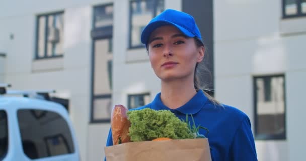 Close up of pretty Caucasian young female delivery worker in blue T-shirt and hat looking with smile at camera and holding packet with goods. Portrait of nice woman with bag full of food outdoors. — Stock Video