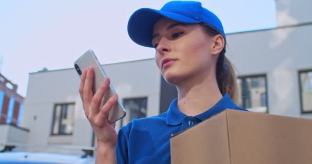 Primer plano de la joven sonriente caucásica bonita mujer en sombrero azul y camiseta sosteniendo paquete y mensaje de texto en el teléfono móvil al aire libre. Atractivo mensajero femenino desplazándose y tocando en el teléfono inteligente . — Vídeos de Stock