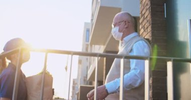 Tıbbi maskeli ve eldivenli beyaz kız, dışarıdaki erkek emekliye taze market alışverişi yaptırıyor. Hizmet konsepti. Coronavirus sırasında yaşlı kadına yiyecek sağlayan dişi kurye..