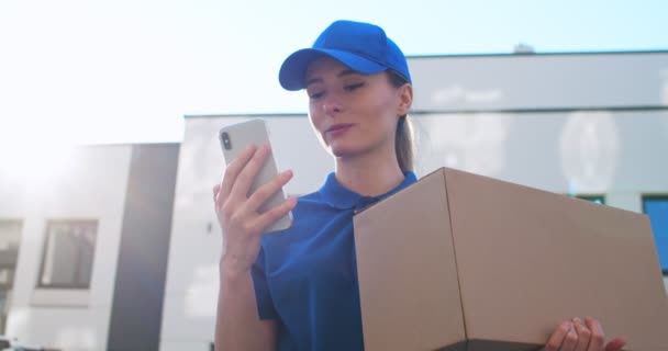 Vista desde abajo en hermosa joven trabajadora postal caucásica en gorra azul y con caja de cartón tocando en el teléfono inteligente al aire libre. Chica bonita mensajes de texto SMS y desplazamiento en el teléfono móvil en la calle . — Vídeos de Stock