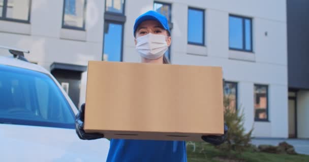 Portrait of young Caucasian beautiful woman in blue uniform, hat and medical mask standing outdoor at street with parcel in hands. Pretty female courier in gloves handing parcel to camera and smiling. — Stock Video