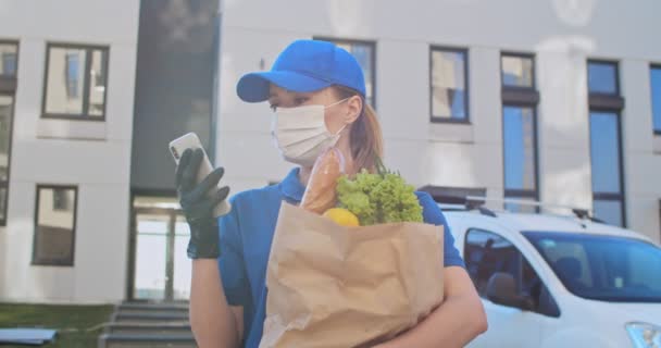Close up of young Caucasian pretty woman courier in uniform, hat, mask and gloves tapping on phone and holding packet with fresh food. Female delivery worker texting and scrolling on smartphone. — Stock Video