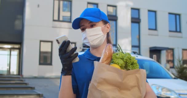 Jonge bezorger vrouw in uniform, hoed, medisch masker, handschoenen tas met verse kruidenier en afluisteren of scrollen op de telefoon buiten. Vrouwelijke koerier sms 'en op smartphone met pakket in handen. — Stockvideo