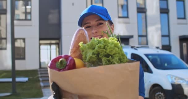 Primer plano de la encantadora joven mujer caucásica, trabajadora de servicio de entrega, en uniforme azul y sombrero sonriendo a la cámara al aire libre con comida fresca en la bolsa. Retrato de la mensajera bastante alegre de la tienda de comestibles — Vídeos de Stock