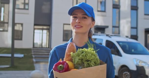 Retrato de atraente jovem caucasiana de uniforme azul e chapéu sorrindo para câmera na rua com comida fresca em pacote. Close-up de muito alegre mulher trabalhador de entrega de mercadorias de supermercado . — Vídeo de Stock