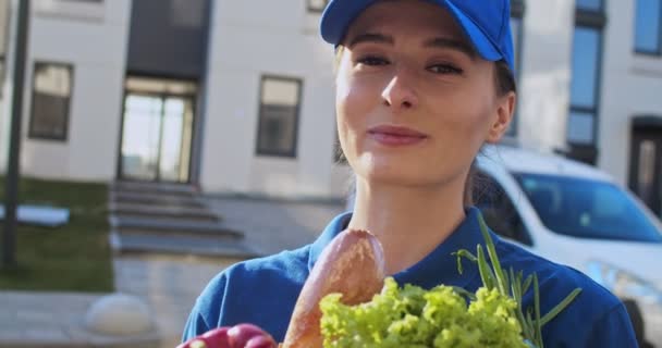 Porträtt av vackra unga kaukasiska kvinna, arbetare av leverans service, i blå uniform och mössa ler mot kameran utomhus med färsk mat i paket. Närbild av ganska glad kvinnlig kurir av livsmedel — Stockvideo