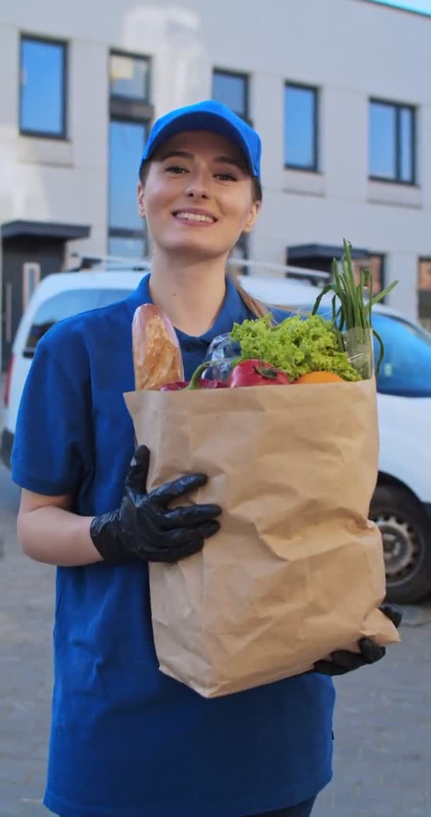 Retrato vertical de la hermosa joven del servicio de entrega, en uniforme azul, guantes y gorra sonriendo a la cámara al aire libre con comida fresca en paquete. Bonita mensajera de abarrotes. Concepto de historias . — Vídeos de Stock