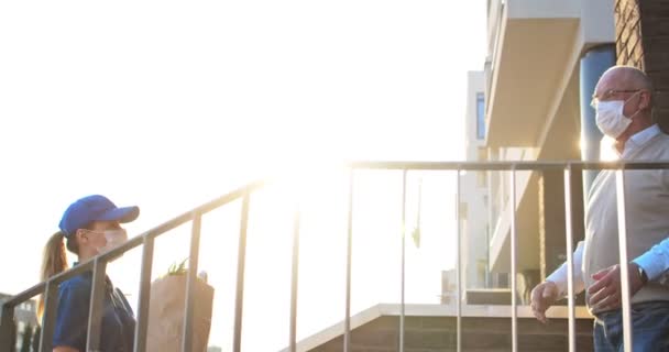 Female courier in medical mask and gloves bringing packet with fresh food to retired old man. Young Caucasian woman handing bag with grocery to male pensioner outdoor at house. Delivery service. — Stock Video