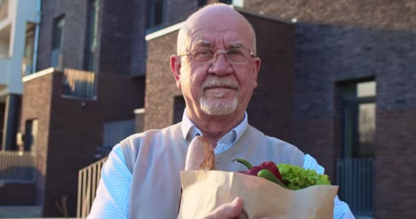 Portrait of Caucasian old retired man in glasses standing outdoor at house, holding packet with fresh food and looking at camera. Market delivery concept. Close up male senior pensioner with grocery — Stock Video