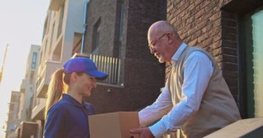 Beyaz bir kız, dışarıda duran erkek emekliye karton kutu getiriyor. Hizmet konsepti. Dişi kurye, gözlüklü yaşlı adama paketi teslim etti ve onunla evin dışında buluştu...