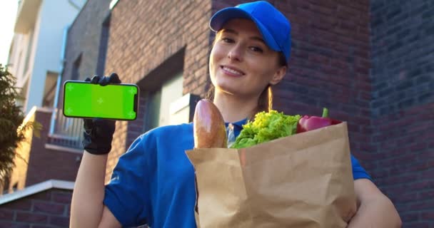 Närbild av vit kvinna ler utomhus och visar horisontell smartphone med grön skärm. Kvinnliga kurir håller paket med mat och demonstrera telefonen. Chroma nyckel. Spårningsrörelse. — Stockvideo