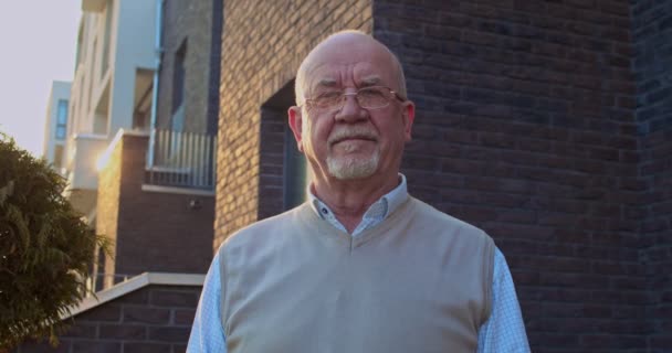 Portrét pohledného starého bělocha v brýlích, stojícího venku u domu a hledícího přímo do kamery. Detailní záběr důchodce v důchodu pózování venku doma. — Stock video