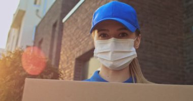 Mavi şapkalı ve tıbbi maskeli genç beyaz bir kadının elinde bir paketle evin dışında kameraya baktığı görüntüleri. Karton kutulu güzel kadın teslimatçının portresi..