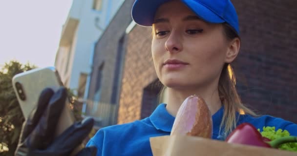 Close up of Caucasian young pretty woman in blue hat and gloves holding food packet and tapping or texting message on smartphone outdoor. Female grocery courier typing and scrolling on phone. — Stock Video