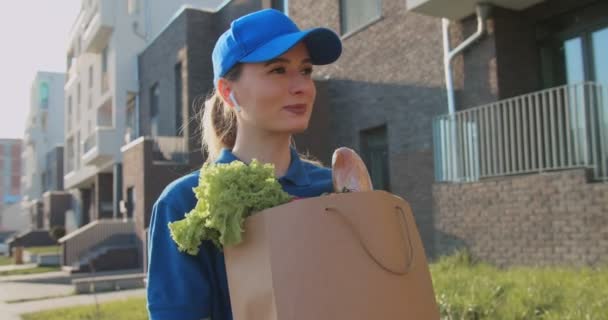 백인젊고 예쁜 여성이고 블루투스 헤드폰 과 모자를 쓰고 거리를 걷고 신선 한 음식이든 봉투를 들고 다니고 있습니다. 시장에서 식료품을 배달하러 가는 아름다운 여급. — 비디오