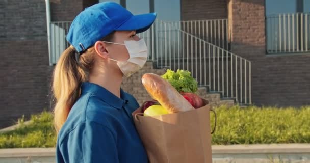 Caucasian young pretty woman, delivery worker in bluetooth headphones, cap and medical mask walking the street and carrying packet with fresh food. Female beautiful courier bringing grocery to house. — Stock Video