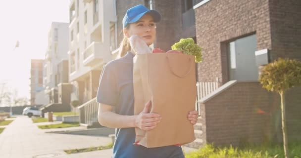 白人の若いかわいい女性、青い制服を着た配達労働者、通りを歩いて新鮮な食べ物でパケットを運ぶキャップ。女性の美しい宅配便をもたらす袋で食料品。コンセプトを伝える. — ストック動画
