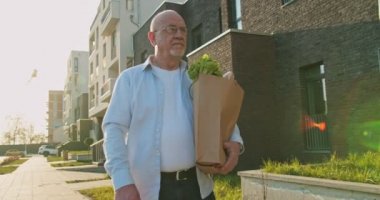 Beyaz gözlüklü yaşlı adam alışveriş yaptıktan sonra sokakta yürüyor ve elinde paketle markete gidiyor. Erkek, emekli emekli, dışarıda yiyecek taşıyan bir emekli. Marketten geliyorum..