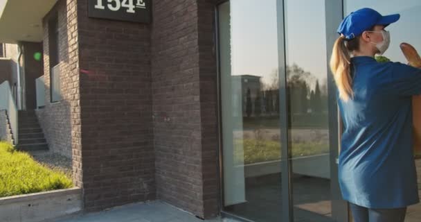 Mujer joven mensajera caucásica en máscara médica trayendo el paquete con la tienda de comestibles al viejo jubilado y entregándolo al aire libre. Mujer con bolsa llena de verduras frescas dándosela a pensionista masculino. Trabajo social — Vídeo de stock