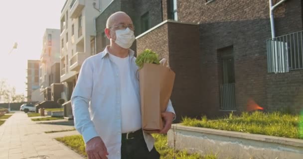 Кавказский старик в очках шел по улице домой после шоппинга и носил пакет с продуктами. Мужчина, пенсионер в медицинской маске, держит еду в сумке на открытом воздухе. Пандемическое время — стоковое видео