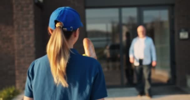 Posteriore di giovane donna caucasica dal servizio di consegna in cappello che porta il pacchetto con il cibo al vecchio in occhiali e lo dà all'aperto. Vista posteriore sul corriere femminile dando generi alimentari consegnare al nonno . — Video Stock