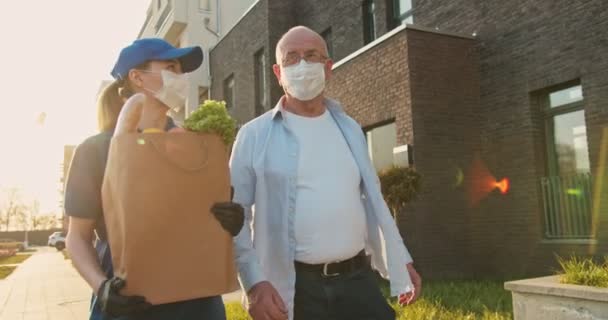 Caucasian young female courier in medical mask, gloves and hat carrying packet with grocery and walking with senior man. Woman from delivering service and male pensioner talking cheerfully outdoor. — Stock Video