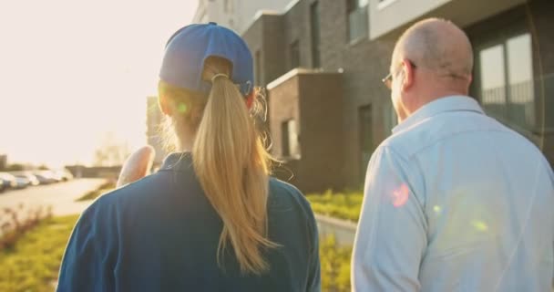 Trasera de mensajera joven caucásica en uniforme llevando paquete con comestibles y caminando con hombre mayor. Vista trasera de la mujer en gorra y pensionista masculino hablando alegremente al aire libre en el paseo . — Vídeos de Stock