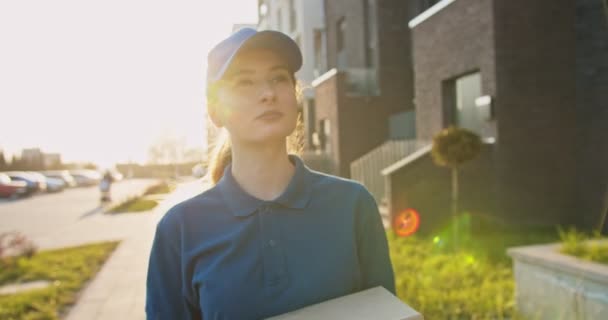 Mujer guapa joven caucásica, trabajadora de reparto en camiseta azul y gorra caminando por la calle y llevando caja de cartón. Mujer hermosa mensajería va a entregar el paquete . — Vídeo de stock