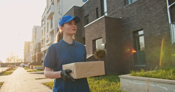 Mujer joven caucásica mensajero entrega caminar y llevar caja de cartón en las manos. Al aire libre. Mujer que lleva paquete de caja de cartón cuando trabaja en el parto. Concepto de correo . — Vídeos de Stock