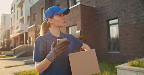 Mujer guapa joven caucásica, trabajadora de reparto en gorra azul caminando por la calle y llevando caja de cartón mientras usa el teléfono inteligente, buscando ruta. Mensajería femenina con paquetes tocando y mensajes de texto en el teléfono . — Vídeos de Stock