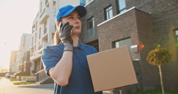 Mujer guapa joven caucásica, trabajadora de reparto en gorra azul caminando por la calle, llevando paquete de caja de cartón y hablando por teléfono móvil. Mujer hermosa mensajería con caja de cartón hablando por teléfono celular . — Vídeos de Stock