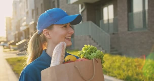 Mujer joven caucásica, trabajadora de reparto en gorra azul caminando por la calle y llevando paquete con comida fresca. Mujer hermosa mensajería que trae la tienda de comestibles del mercado al hombre mayor . — Vídeo de stock