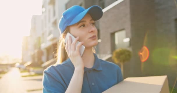 Zbliżenie na ładną młodą białą kobietę w niebieskim kapeluszu rozmawiającą przez telefon komórkowy z uśmiechem podczas spaceru ulicą w słoneczny dzień. Kurierka z paczką kartonową rozmawiająca przez telefon komórkowy na zewnątrz. — Wideo stockowe