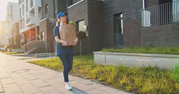 Mulher branca bonita jovem em chapéu azul e camiseta andando na rua no dia ensolarado e carregando pacote com comida. Correio feminino com saco de supermercado nas mãos entregando-o. Ao ar livre . — Vídeo de Stock