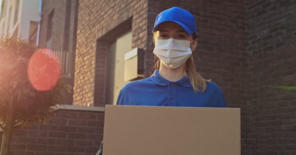 Retrato de la joven mujer blanca hermosa en uniforme azul, sombrero y máscara médica de pie al aire libre en la casa con paquete en las manos. Bastante mujer mensajero entrega caja de cartón a la cámara . — Vídeos de Stock