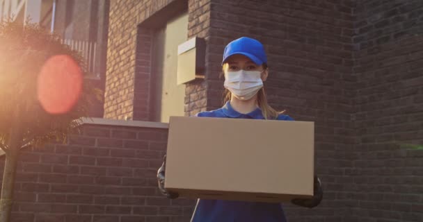 Retrato de la joven mujer caucásica feliz en uniforme azul, gorra y máscara médica de pie al aire libre en la casa con el paquete en las manos y sonriendo. Bastante trabajadora de reparto entregando caja a cámara . — Vídeos de Stock