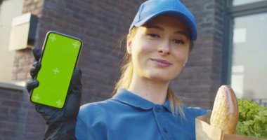 Eldivenli beyaz kadın portresi. Elinde yeşil ekranlı dikey akıllı telefon gösteren bir paket yiyecekle gülümsüyor. Dışarıdaki kadın kurye, elinde sebzelerle telefon gösterisi yapıyor. Krom anahtar. İzleme hareketi