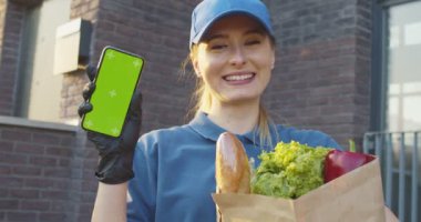 Açık havada gülümseyen ve dikey akıllı telefonu yeşil ekranlı gösteren beyaz bir kadın. Dişi kurye pakette yiyecek ve telefon gösterisi yapıyor. Krom anahtar. İzleme hareketi.