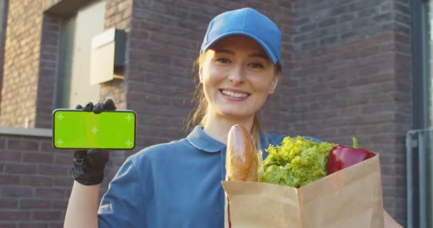 Zbliżenie kaukaska kobieta uśmiecha się na zewnątrz i pokazuje poziomego smartfona z zielonym ekranem. Kurierka trzymająca paczkę z jedzeniem i demonstrująca telefon. Klucz chromowy. Ruch śledzenia. — Wideo stockowe