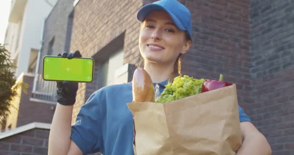 Portrait of Caucasian woman in gloves smiling with packet of food showing horizontal smartphone green screen. Female courier outdoor with vegetables demonstrating phone. Chroma key. Tracking motion. — Stock Video