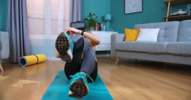 Giovane donna caucasica sportiva che indossa occhiali di realtà virtuale mentre si siede sul tappetino yoga e si allena. Adatta all'allenamento femminile, all'allenamento e al fare esercizi addominali flettendo le gambe sul pavimento a casa, vista frontale . — Video Stock